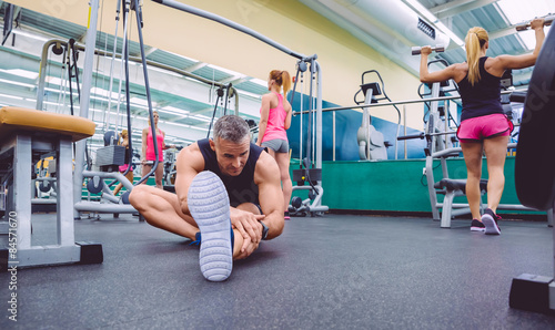 Fototapeta Man stretching and women doing dumbbells exercises in gym