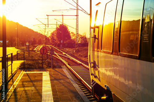 Plakat Modern passenger train standing at countryside platform with beautiful landscape at sunset.